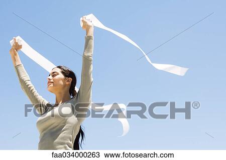Stock Image - Woman holding up rolls of paper outdoors, eyes closed, low angle view. Fotosearch
