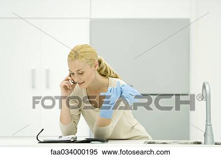Woman in rubber gloves leaning against kitchen counter, using cell phone, looking at agenda View Large Photo Image Stock Photography - Woman in rubber gloves leaning against kitchen counter, using cell phone, looking at agenda. Fotosearch