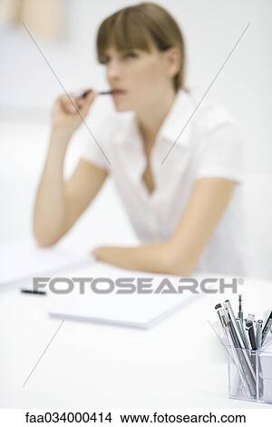 Woman seated at desk, pen in mouth, focus on container full of pens in foreground View Large Photo Image Picture - Woman seated at desk, pen in mouth, focus on container full of pens in foreground. Fotosearch