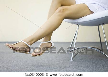 Stock Image - Woman sitting in chair with legs dangling, wearing sandals, cropped view. Fotosearch