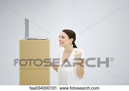 Stock Image - Woman smiling with hands raised, looking at laptop computer set high on cardboard box. Fotosearch