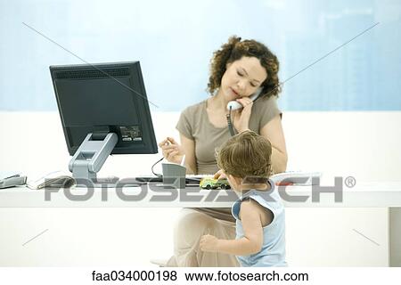 Stock Photo - Woman using phone in office, toddler son playing with toy truck on desk. Fotosearch
