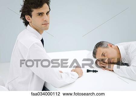 Stock Image - Young businessman sitting across table from defeated older businessman, chess piece between them. Fotosearch