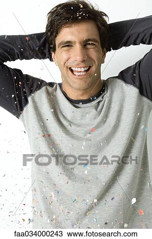 Stock Image - Young man amidst falling confetti, laughing at camera, hands behind head. Fotosearch