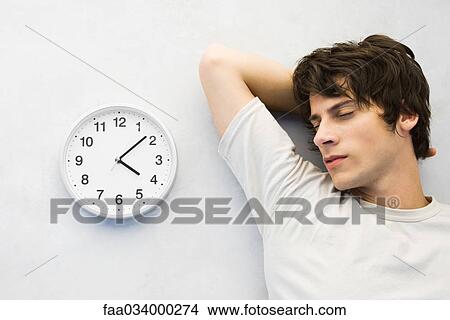 Picture - Young man leaning against wall beside clock, eyes closed. Fotosearch