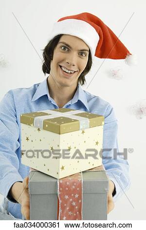Young man wearing Santa hat, holding out Christmas gifts, smiling at camera View Large Photo Image Stock Image - Young man wearing Santa hat, holding out Christmas gifts, smiling at camera. Fotosearch
