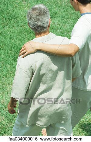 Young man with his arm across the shoulder of an older man walking together across grass View Large Photo Image Stock Photograph - Young man with his arm across the shoulder of an older man walking together across grass. Fotosearch