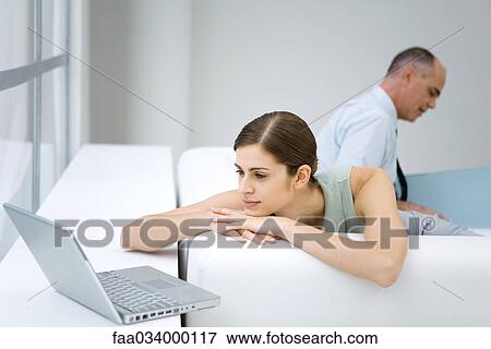 Stock Photo - Young woman relaxing on sofa, using laptop computer, businessman reading in background. Fotosearch
