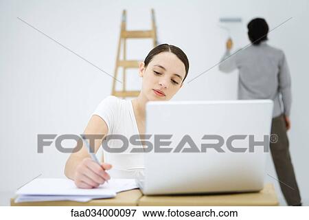 Stock Photo - Young woman using laptop computer, man in background painting wall. Fotosearch