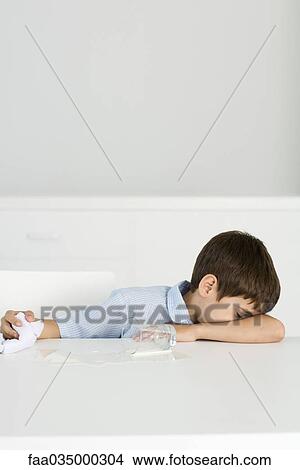 Picture - Boy sitting at table with spilled milk, head down, holding towel. Fotosearch