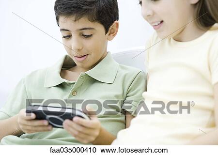 Stock Image - Brother and sister sitting together, boy playing handheld video game. Fotosearch