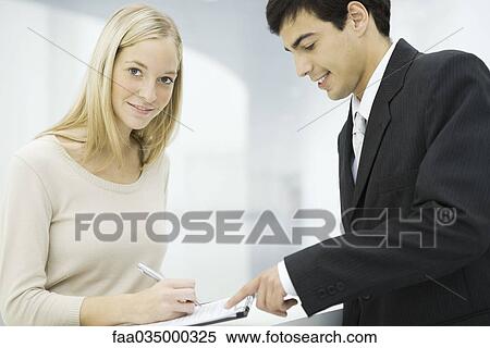 Businessman holding out clipboard, woman writing on document and smiling at camera View Large Photo Image Stock Photography - Businessman holding out clipboard, woman writing on document and smiling at camera. Fotosearch