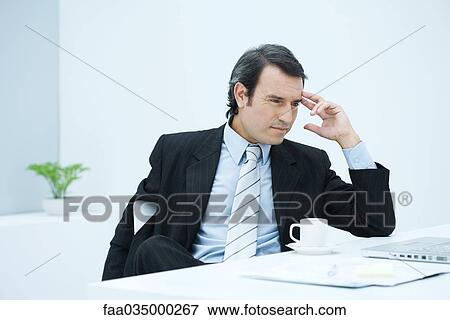 Stock Photo - Businessman sitting at desk, holding head, looking away. Fotosearch