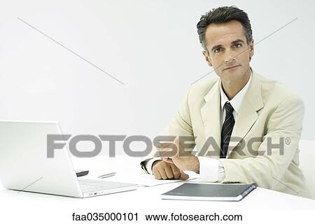 Stock Image - Businessman sitting at desk, looking at camera. Fotosearch