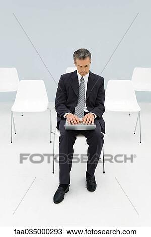 Businessman sitting in chair using laptop, empty chairs in background View Large Photo Image Stock Image - Businessman sitting in chair using laptop, empty chairs in background. Fotosearch