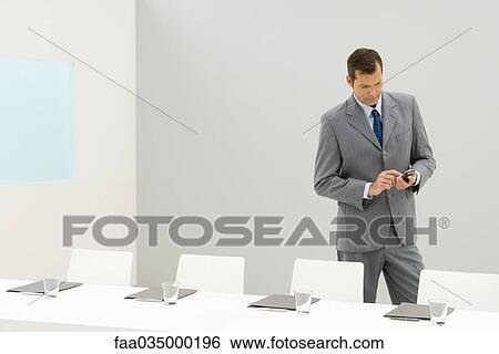 Stock Photograph - Businessman standing beside conference table, looking down at cell phone. Fotosearch