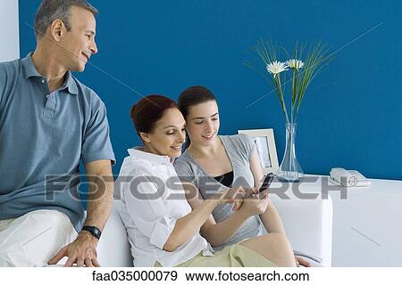 Stock Photo - Family sitting on couch, teen daughter showing mother cell phone, father watching. Fotosearch