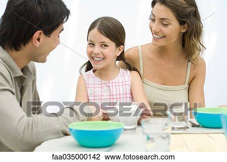 Stock Image - Family sitting together at the table, smiling at each other. Fotosearch