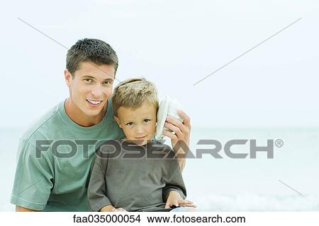 Picture - Father holding seashell up to son's ear, both smiling at camera. Fotosearch