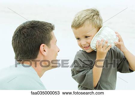 Stock Image - Father with son at beach, boy listening to seashell. Fotosearch