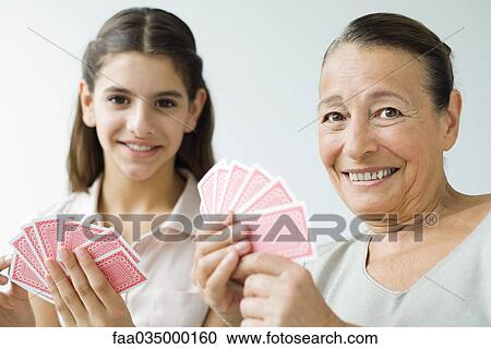 Stock Image - Grandmother and granddaughter playing cards, smiling at camera. Fotosearch