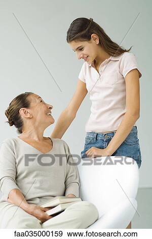 Stock Photo - Grandmother sitting in chair, holding book, smiling up at teen granddaughter standing nearby. Fotosearch