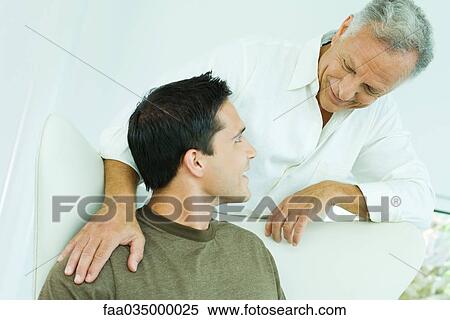 Stock Photography - Man leaning over adult son's shoulder, both smiling at each other. Fotosearch