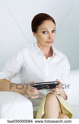 Picture - Mature woman holding handheld video game, looking at camera with eyebrow raised. Fotosearch