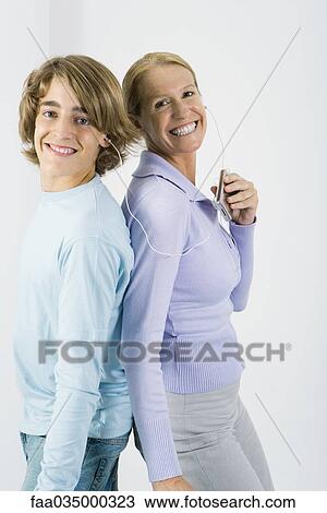 Stock Image - Mother and teen son listening to MP3 player together, standing back to back, smiling at camera. Fotosearch