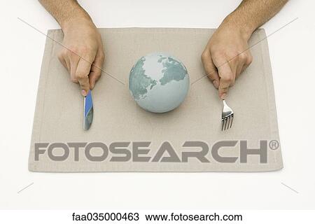 Stock Image - Person sitting before globe on placemat, holding fork and knife, cropped view. Fotosearch