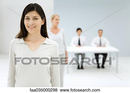 Stock Photo - Professional woman smiling at camera, colleagues in background. Fotosearch