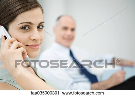 Professional woman using cell phone, smiling at camera, male colleague in background View Large Photo Image Stock Image - Professional woman using cell phone, smiling at camera, male colleague in background. Fotosearch