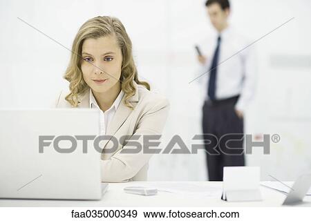 Stock Photo - Professional woman using laptop computer at desk, man standing in background. Fotosearch