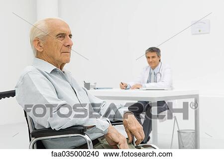 Stock Image - Senior patient sitting in wheelchair, doctor working at desk in background. Fotosearch
