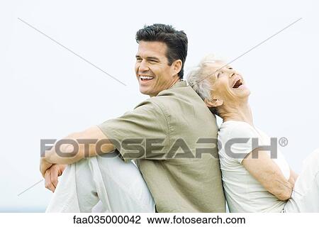 Stock Image - Senior woman and adult son sitting back to back, laughing, side view. Fotosearch