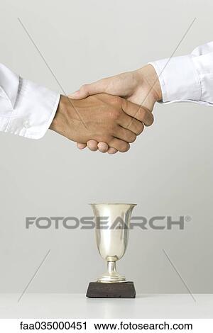 Stock Image - Shaking hands over trophy, cropped view. Fotosearch