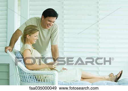 Stock Photo - Woman relaxing in chair, reading book, man leaning over her shoulder and smiling. Fotosearch