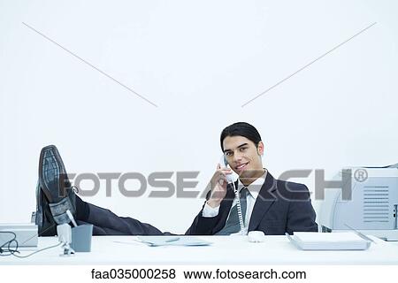 Young businessman sitting at desk with feet up, talking on landline phone View Large Photo Image Stock Photo - Young businessman sitting at desk with feet up, talking on landline phone. Fotosearch