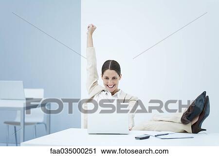 Stock Image - Young businesswoman sitting at desk, smiling at laptop and raising one arm in the air. Fotosearch