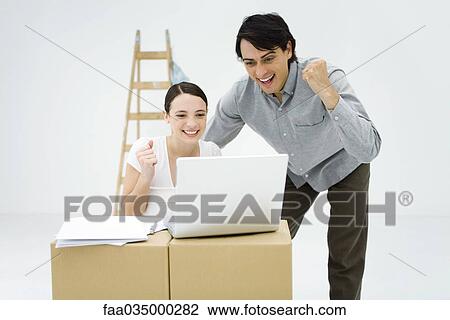 Young couple looking at laptop computer on top of cardboard box, smiling, clenching fists View Large Photo Image Stock Image - Young couple looking at laptop computer on top of cardboard box, smiling, clenching fists. Fotosearch