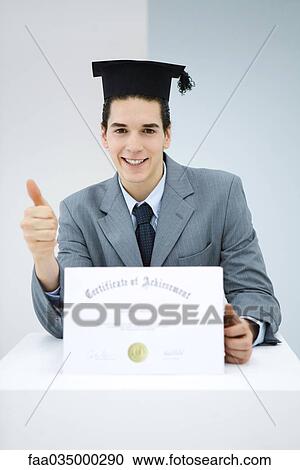 Stock Image - Young man with diploma, wearing graduation cap, making thumbs-up gesture. Fotosearch