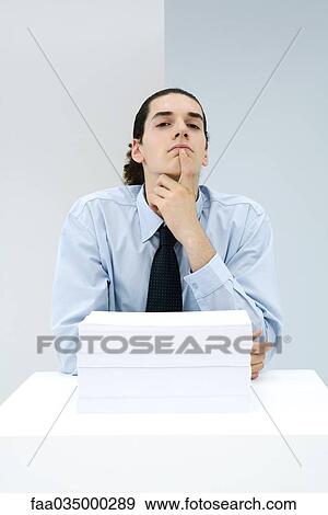 Stock Photo - Young office worker behind stack of papers, hand under chin, looking at camera. Fotosearch