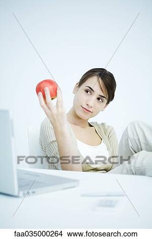 Picture - Young woman sitting at desk, looking at apple in hand. Fotosearch