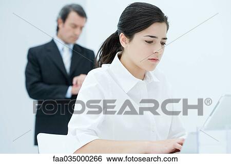 Stock Photograph - Young woman typing on laptop, boss checking time in background. Fotosearch