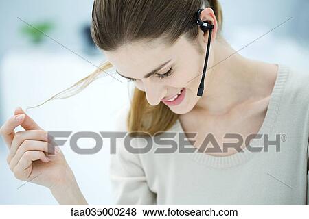 Stock Photo - Young woman wearing headset, playing with hair and looking down, smiling. Fotosearch