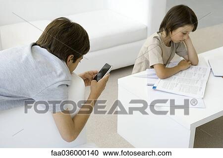 Boy reading at coffee table, brother leaning on armchair, holding cell phone View Large Photo Image Stock Image - Boy reading at coffee table, brother leaning on armchair, holding cell phone. Fotosearch