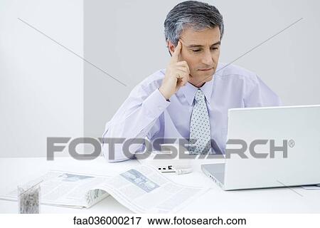 Businessman sitting at desk, looking at laptop computer, holding head View Large Photo Image Stock Photo - Businessman sitting at desk, looking at laptop computer, holding head. Fotosearch
