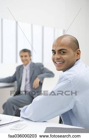 Businessman sitting at desk, smiling over shoulder at camera, colleague in background View Large Photo Image Stock Image - Businessman sitting at desk, smiling over shoulder at camera, colleague in background. Fotosearch