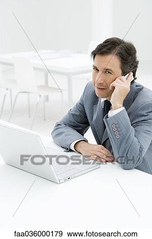 Businessman talking on cell phone, sitting by laptop computer View Large Photo Image Stock Photo - Businessman talking on cell phone, sitting by laptop computer. Fotosearch