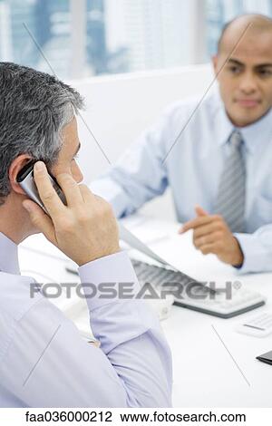 Stock Image - Businessmen having a meeting at desk, man in foreground talking on cell phone. Fotosearch